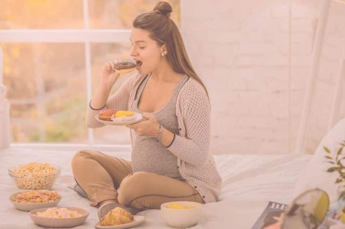 Beitragsbild Blogartikel Ernährung in der Schwangerschaft - Schwangere Frau sitzt zwischen Essen und beißt in einen Donut